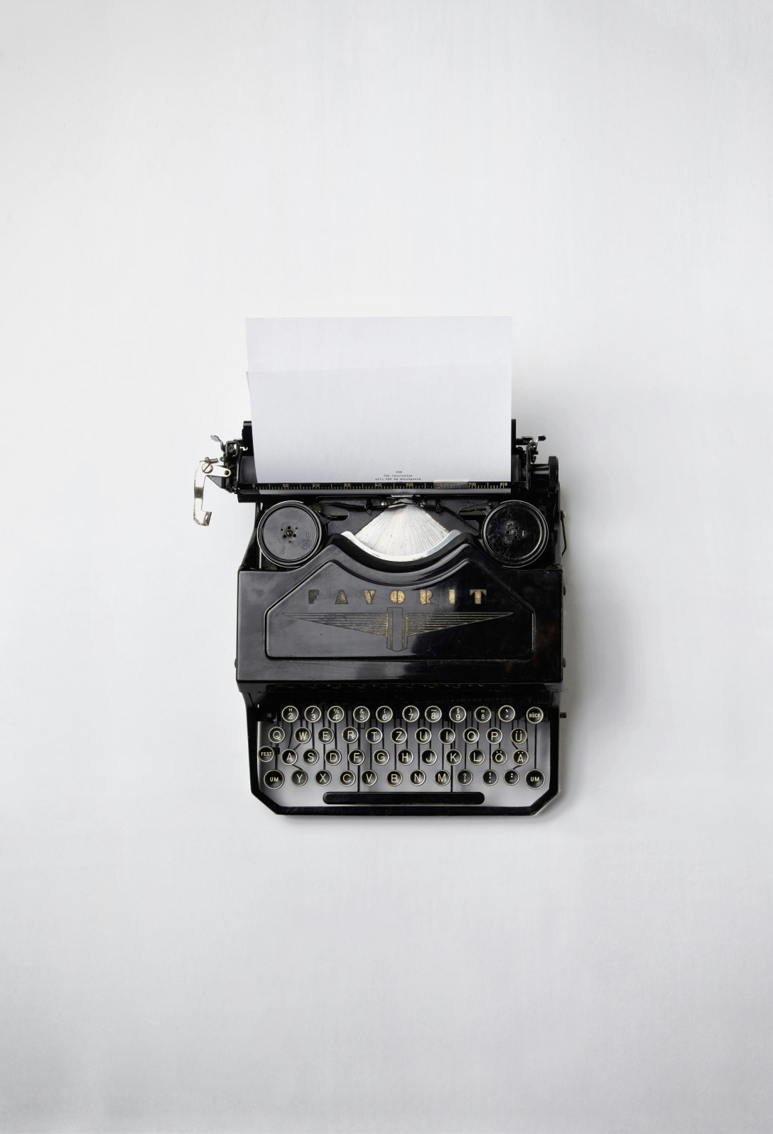 A minimalist photograph of a vintage black typewriter with a blank sheet of paper inserted, centred against a clean white background, evoking reflection, writing, and the quiet act of putting words to page.